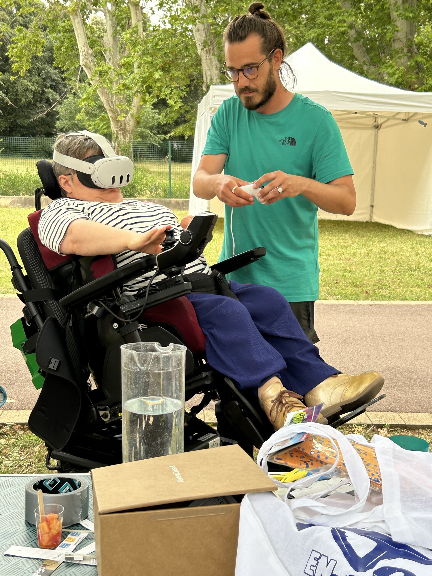 Une personne sur un fauteuil roulant avec un casque virtuel et un animateur en face qui règle le matériel.